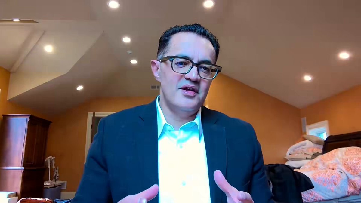 The image shows a man wearing glasses, sitting at a desk with his hands outstretched as if he's giving a presentation or speech. The background features an orange wall that complements the man's attire. There are also two other people visible in the scene, one on the left side and another person partially hidden behind the man. The image is related to the post because it contains text about a man giving a presentation at Monument Bank, which could be interpreted as him being interviewed or discussing financial matters.