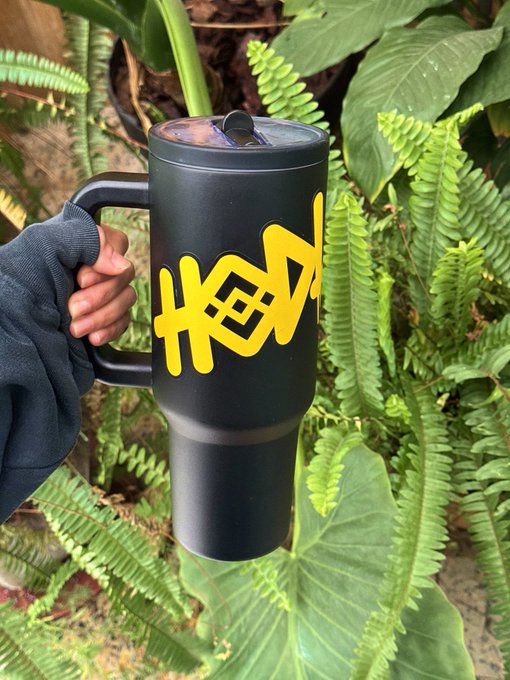 A hand clad in a dark sleeve holds up a tall black tumbler featuring bold yellow "HODL" graffiti-style lettering against a lush backdrop of green ferns and foliage, perfectly capturing the weekend's relaxed crypto "HODLing" vibe.