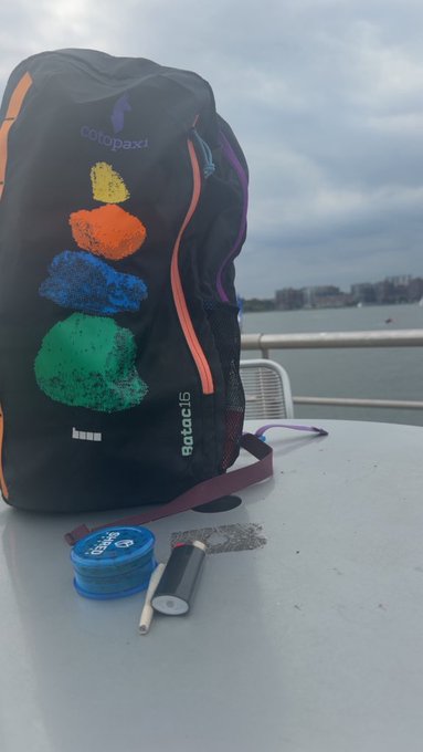 A black Cotopaxi 'gotac15' backpack with colorful stacked blob graphics sits on a table next to a blue container and earbuds, with water in the background.