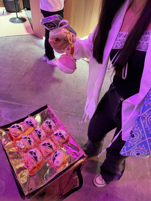 A person holds up a small pink plush keychain at what appears to be a fan event, with a display of orange-wrapped bear merchandise visible in a lit case below.