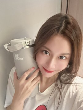 A young woman with long brown hair holding a white MAHOTSUKU branded water bottle close to her face.