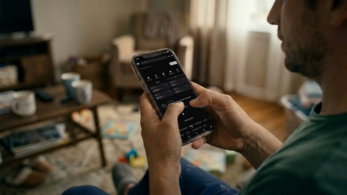 The image shows a man sitting down, holding a smartphone that has an app open displaying a map of his location. The man appears to be using the phone for navigation or planning purposes. The background reveals a living room setting with various objects such as a couch, chair, and books scattered around.