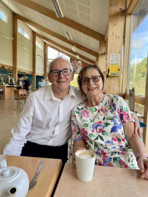 An older couple, a man in a white shirt and a woman in a floral dress, smile together while seated at a table with a teapot and coffee mug in a brightly lit room with large windows.