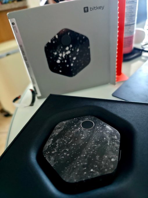 A Bitkey hardware wallet is displayed next to its hexagonal packaging, showcasing the new bitcoin hardware device's sleek design and matching box art. The image visually confirms the user's recent acquisition of this Bitcoin cold storage device, which they describe as having solid build quality, reliable NFC functionality, and smooth onboarding—though with occasional cloud storage errors likely related to their VPN connection.