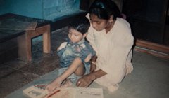 An indoor scene shows a woman in a white shirt helping a small child in a blue outfit sit on the floor next to a wooden stool.