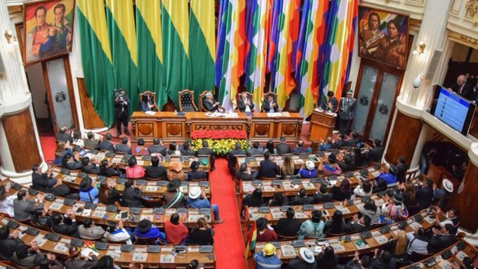 A legislative chamber filled with seated officials, colorful flags, and portraits on the walls, appearing to be the Bolivian parliament.