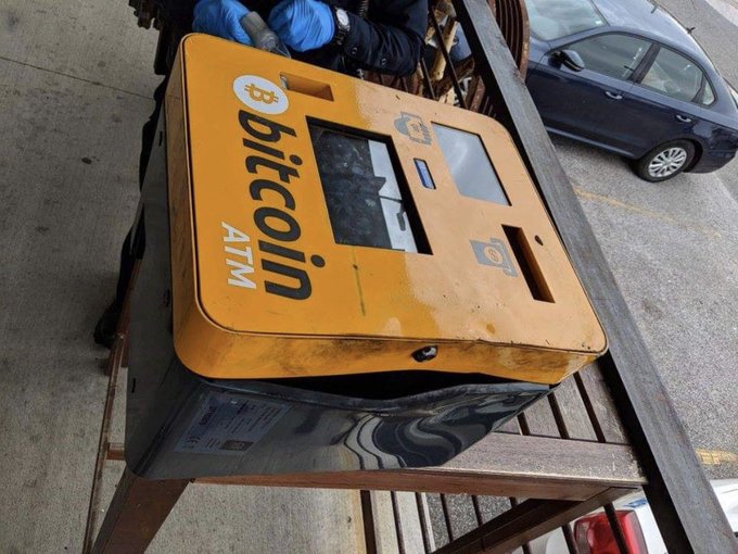 An orange Bitcoin ATM machine mounted on a wooden structure, with a person wearing blue gloves working on it, and a car parked in the background.