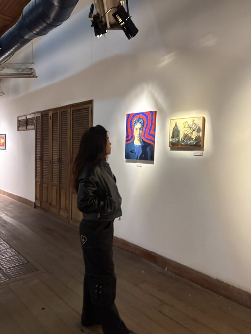 A person with long dark hair wearing a leather jacket stands in a gallery viewing colorful paintings on a white wall.