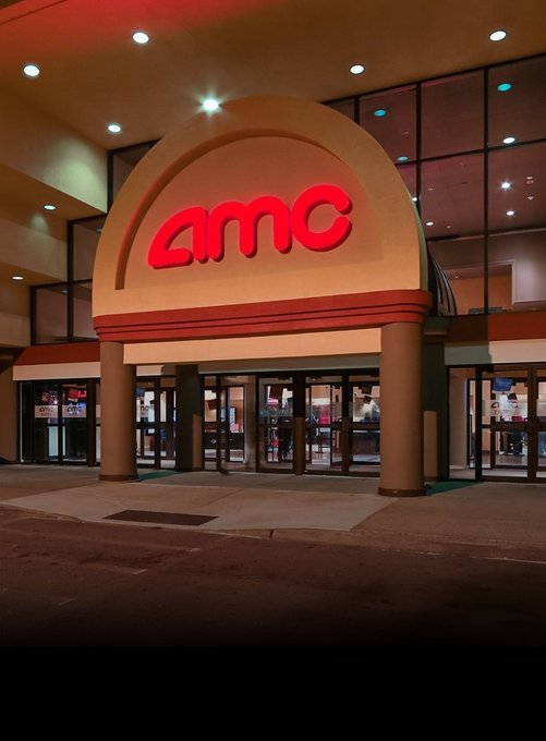 The illuminated AMC Theatres entrance features the bold red "amc" logo on a tan archway, with glass doors below. The image supports the post's announcement that AMC theaters now accept XRP as a payment method through BitPay.