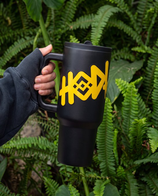 A hand in a dark sleeve holds a black insulated travel mug featuring bold yellow "HODL" lettering, set against a lush backdrop of green ferns—visually reinforcing the "caffeine to hodl the day" theme by pairing crypto culture with a rugged, ready-for-anything vibe.