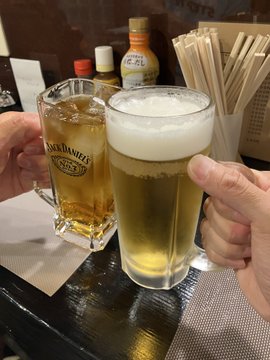 Two hands clink glasses of cold beer—one a mug branded "JACK DANIEL'S #7" and the other a tall pilsner glass filled to a frothy head—at a bar with chopsticks in the background. The toast appears to celebrate a stop in Shiraoi or Chitose during a trip geared toward carbo-loading for upcoming races like the Saroma Lake Ultramarathon and Mt. Fuji Hill Climb.