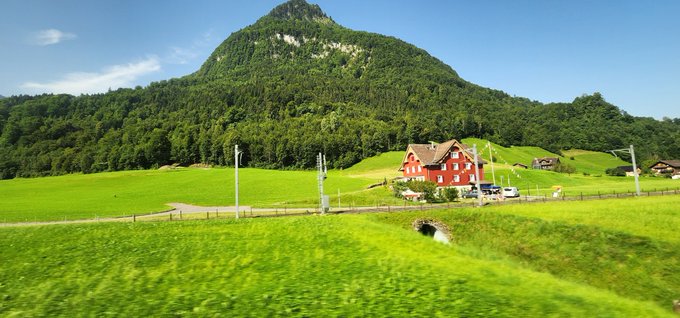 A scenic view of a lush green mountain and farmland featuring a red farmhouse, likely in Switzerland, viewed from a moving train.