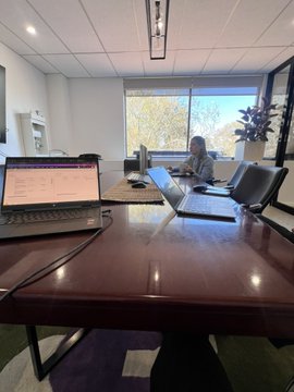 A person sits at a desk with a laptop and a monitor displaying code or data in a modern office with large windows and views of trees.