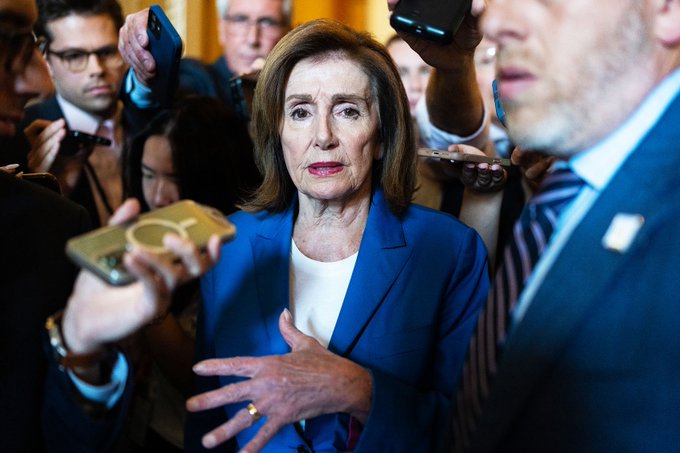 Nancy Pelosi stands centrally surrounded by a crowd of people holding up smartphones, gesturing with her hand as if addressing the media.