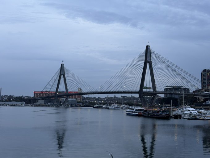 An overcast view of the Anzac Bridge in Sydney, Australia, with a red train crossing the cable-stayed bridge over calm harbor waters, with boats docked in the foreground.