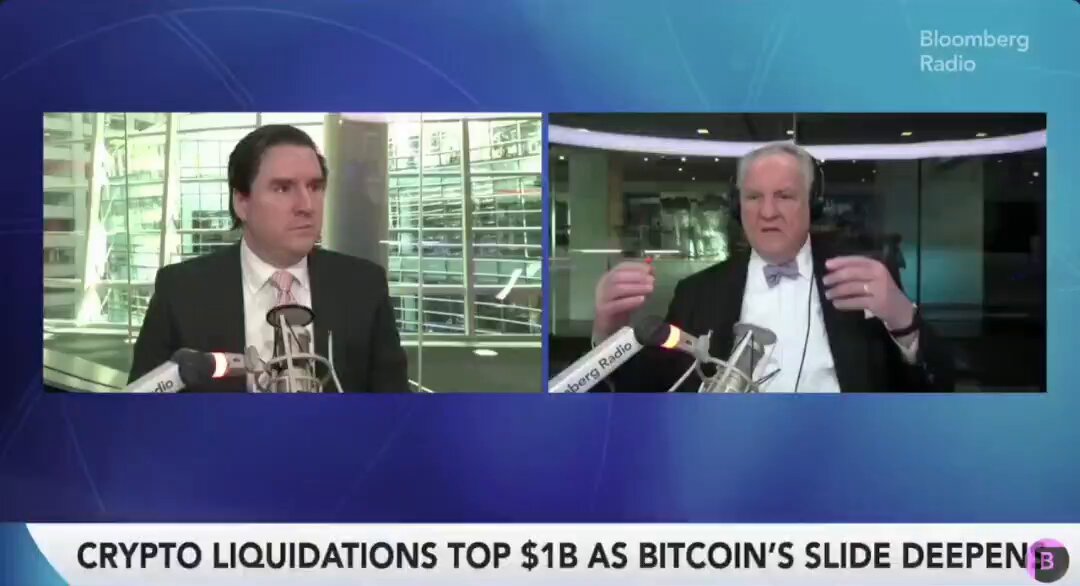 The image shows two men wearing suits, one of whom is holding a microphone while talking about Bitcoin. The other man has his hand raised with a coin attached to it. The background features a blue screen displaying the text "Crypto Liquidations Top 18 As BitCoin's Slide Deepen B". This suggests that the conversation might be related to cryptocurrency trading or investing in Bitcoin, as they are discussing its potential value and market trends.
