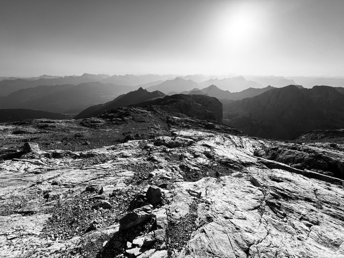 A black-and-white photograph of a rocky mountain landscape with the sun shining brightly in the sky, showcasing layers of distant mountain ranges fading into the horizon.