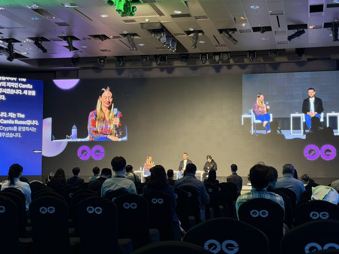Conference event with audience members facing a stage where two panelists are seated; large screens display Korean text about The Camila and crypto investments, with "OG" logos on the chairs.