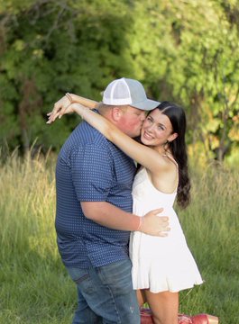A couple embraces outdoors in a grassy field with trees in the background, the man wearing a blue checkered shirt and gray cap, the woman in a white dress.