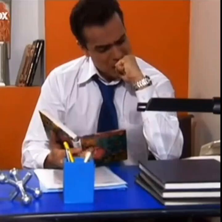 The image shows a man sitting at a desk with a book open in front of him. The desk also has several books, papers, and other items scattered around. There are two blue cups placed near the edge of the desk, as well as a pen holder filled with pens. A clock is visible on the wall behind the man, indicating the time while he reads his book or works at his desk.