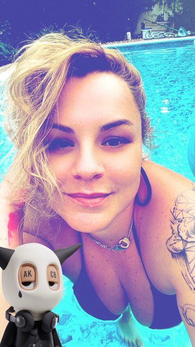 A blonde woman smiling in a swimming pool, with a small cartoon figurine wearing a mask labeled 'AK CS' in the foreground.