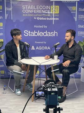 The image shows two men sitting at a table, engaged in a conversation. They are positioned behind a camera that is set up to record their discussion. The backdrop of the scene features a blue wall with white text, which reads "Back in CDMX for the @bitso Stablecoin Conf. LatAm is at the center of drawing the builders, operators, and infrastructure teams that are moving stablecoins forward Building on $AUSD momentum in the region is real Stay tuned for @wensingfacundo 's conversation with @StableDash". The men appear to be discussing a topic related to stablecoins or cryptocurrency.