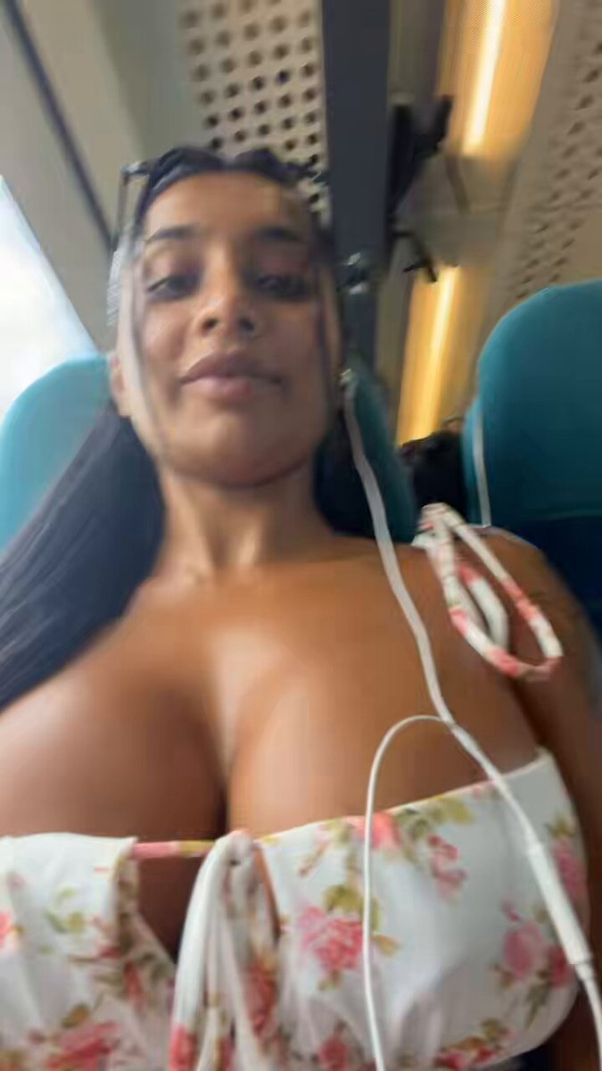 The image features a woman with large breasts wearing a white top, sitting inside a train car. She has her hair pulled back into a ponytail and is looking directly at the camera. The background of the photo shows the interior of the train car, which includes seats and windows. There are also headphones visible in the scene, suggesting that she might be listening to music or watching something on her phone during the ride.