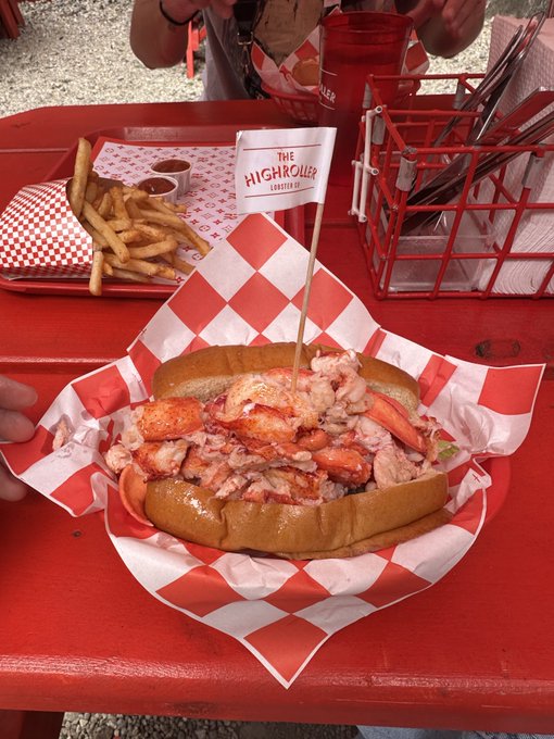 A lobster roll served in a red-and-white checkered paper basket sits on a red tray alongside fries, marked with a 'The Highroller' pick flag.
