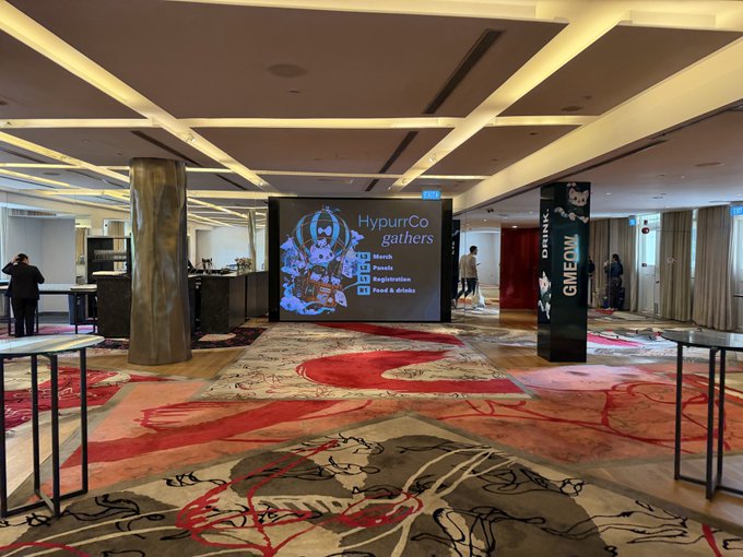 A spacious indoor lobby or lounge area with patterned red and beige carpet, decorative ceiling beams, and a large digital screen displaying "HypurCo gathers" with food and drink icons (Munch, Fuzzle, Nocturnal, Food & Drinks).