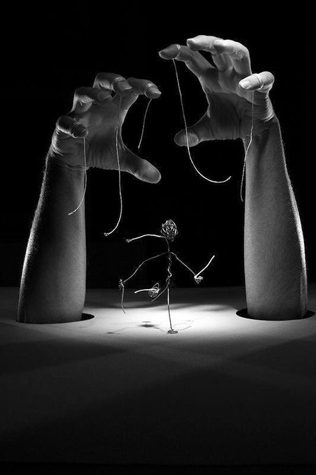 A black and white surrealist photograph showing two large disembodied hands emerging from a surface, manipulating strings attached to a small wire figurine like a marionette puppet.