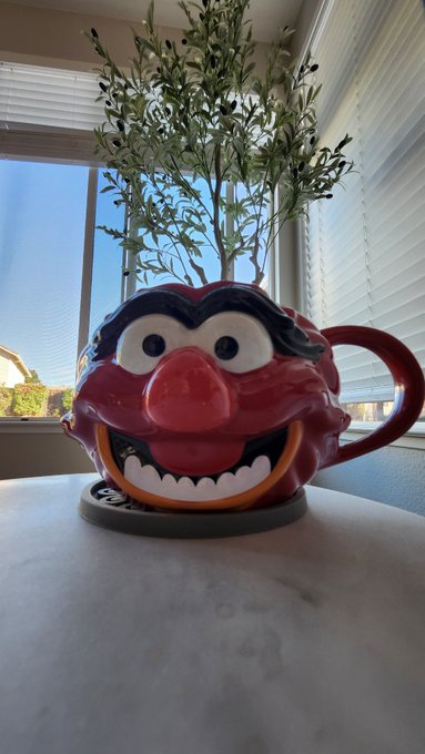 A red novelty coffee mug designed to look like Animal from The Muppets, featuring his face with eyes, nose, and a wide grin, holding a small olive plant on a marble surface.