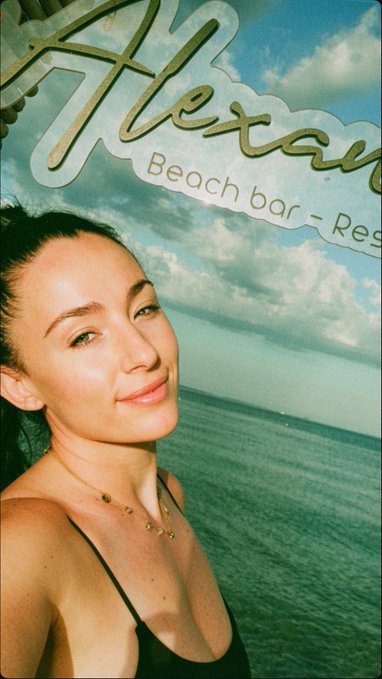 A selfie of a smiling woman with dark hair and a gold necklace, taken in front of a sign for 'Hexagon Beach bar - Res[taurant]' with the ocean and cloudy sky in the background.