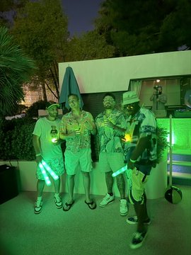 Four friends in summer attire pose together at what appears to be an outdoor evening event, illuminated by green lighting and holding glowing green lightsabers that visually echo the brand color of XRP cryptocurrency. The image ties to the post's mention of an "XRP fam" gathering in Vegas, suggesting the group is part of the cryptocurrency's community celebrating at a meet-up.