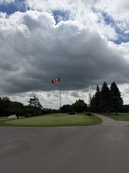 A Canadian flag flying on a flagpole against a dramatic cloudy sky, with a green golf course and trees visible in the background.
