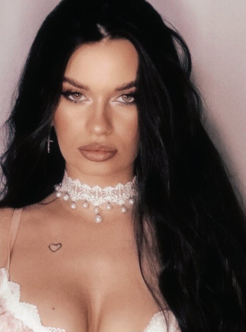 A woman with long dark hair, dramatic eye makeup, a small heart chest tattoo, a cross earring, and a pearl lace choker poses for a portrait.