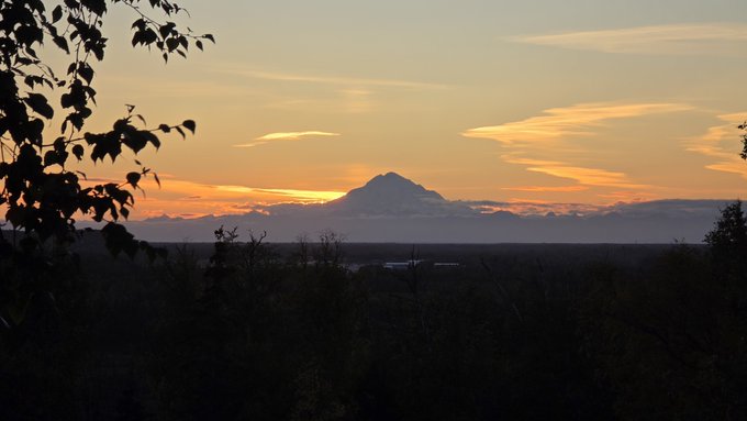 A landscape photo captures a sunset or sunrise over a body of water, with a prominent mountain peak silhouetted in the distance and tree branches framing the left side of the shot.