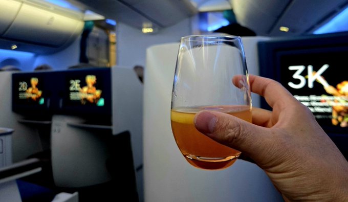 A hand raises a glass of what appears to be white wine or champagne inside a luxury airplane cabin, with seat numbers "2D," "2E," and "3K" visible on the seatback screens in the background. The image captures the "FirstClass" experience being celebrated in the post, showcasing the lavish travel lifestyle funded by crypto profits.