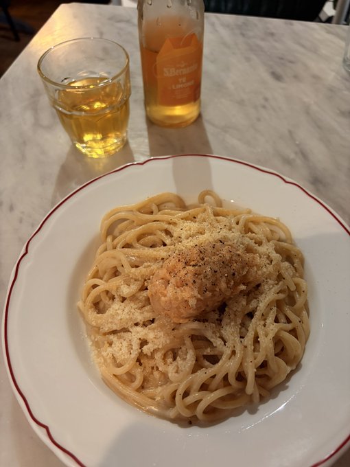 A plate of spaghetti topped with grated cheese and black pepper sits on a marble table, accompanied by a glass of orange beverage and a bottle labeled 'Sikensey'.