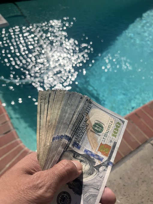 A hand fanning out a thick bundle of $100 US bills by a sunlit swimming pool, with shimmering turquoise water and red brick coping visible in the background.