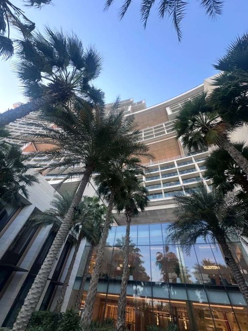 Tall palm trees frame a luxurious outdoor shopping and hotel complex under a clear blue sky, with a Valetini storefront visible at the entrance. The high-end architecture reflects an aspirational, money-focused lifestyle that complements the post's "GM (get money)" message.