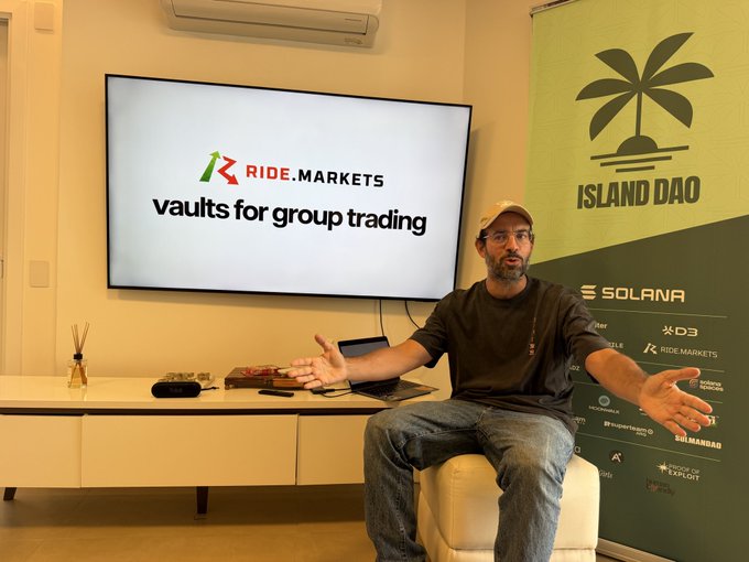 A man sits in an office speaking with open hands beside a screen displaying “RIDE.MARKETS” and “vaults for group trading,” while an “ISLAND DAO” banner and “SOLANA” logos are visible behind him.
