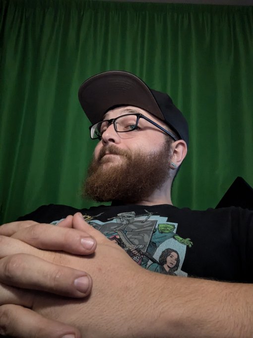 A bearded man wearing glasses and a baseball cap backwards, with his arms crossed, posing in front of a green curtain backdrop.