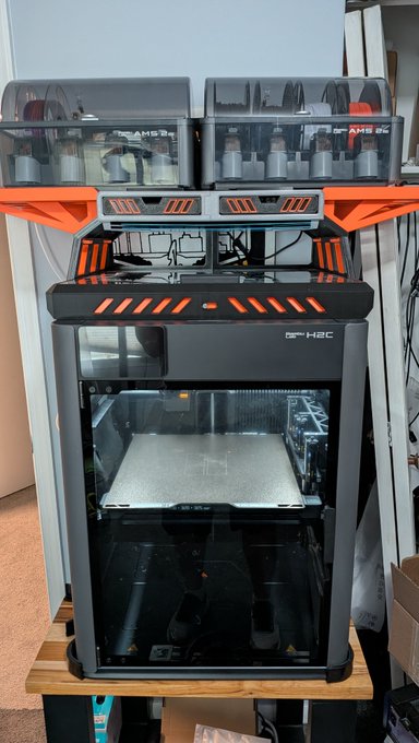 A black and orange 3D printer with AMS (Automatic Material System) filament hoppers mounted on top, sitting on a wooden table.