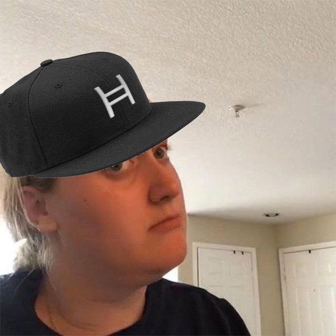 A low-angle selfie of a person wearing a black snapback cap with a white 'H' logo, photographed indoors against a white ceiling.