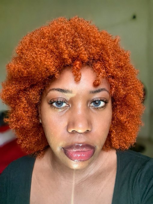 A selfie of a woman with vibrant orange curly afro hair and light-colored eyes, showing some product on her face.