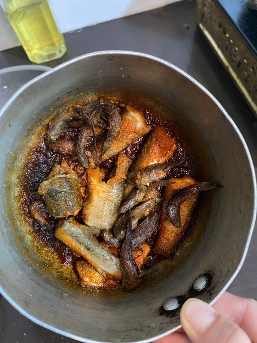 A metal bowl filled with a cooked dish of fish and meat pieces in a dark, rich sauce, with cooking oil visible in the background.