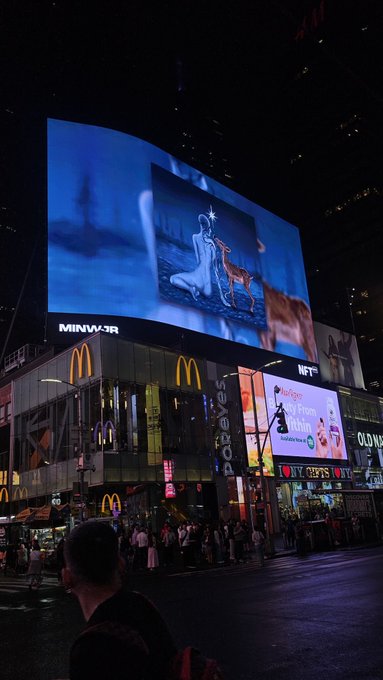 A nighttime street scene in a busy commercial district (appears to be Times Square area) with a large billboard displaying an artistic image, alongside McDonald's and Forever 21 storefronts.