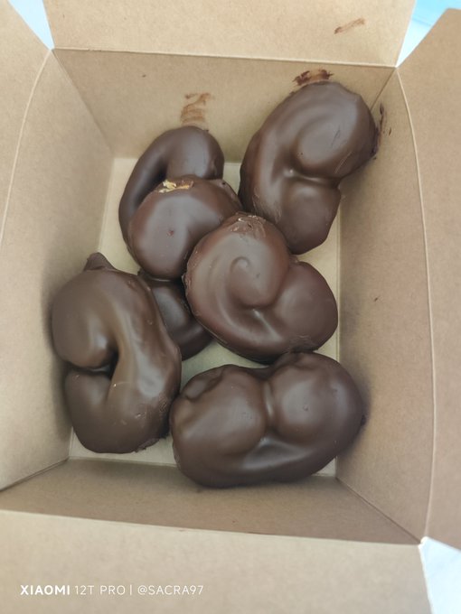 A cardboard box contains several chocolate-covered treats with an irregular, rounded shape, photographed with a 'XIAOMI 12T PRO | @SACRA97' watermark in the bottom left corner.
