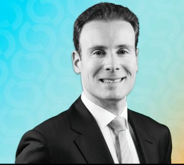 A black-and-white headshot of a smiling middle-aged man in a suit and tie, set against a light blue patterned background.