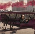 A low-resolution image shows a pilot seated in the open cockpit of an old red aircraft, with smoke and city buildings in the background.